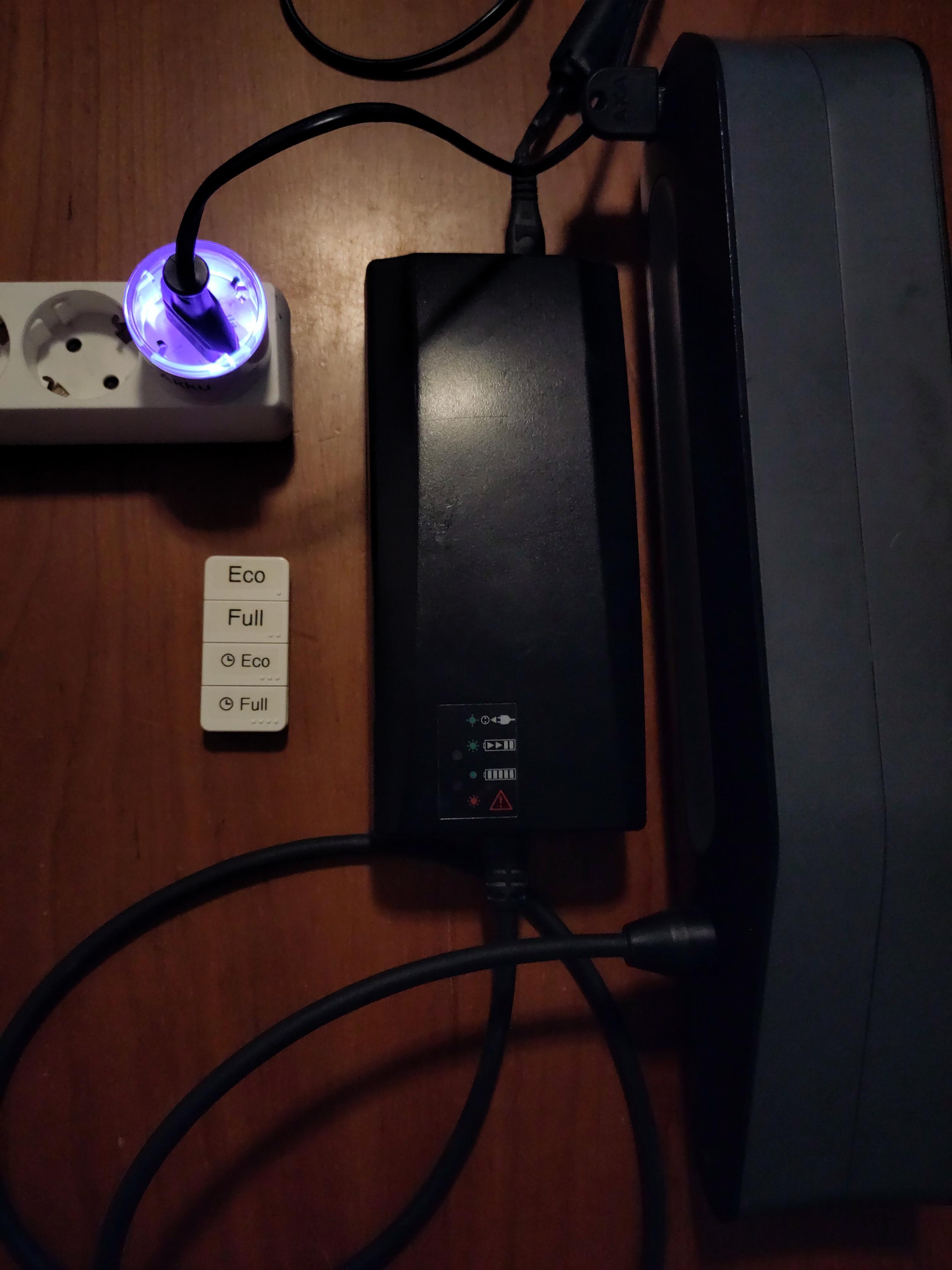 Shelly Plug mit Ladekontroll Software steckt in Steckdosenleiste. Daneben E-Bike Ladegerät und Akku. Ausserdem eine kleine 4-Tasten Bluetooth Fernbedienung mit der Beschriftung Eco und Full und dann noch mal Tasten mit einem Uhr-Symbol und Eco/Full.